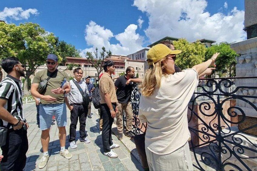 A group of people standing on a paved area with a metal fence, trees, and buildings in the background. 1