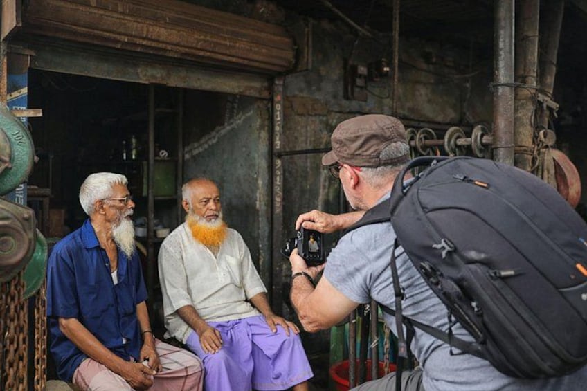 Trois hommes sont assis, l’un d’eux tient un appareil photo. Ils se trouvent dans un cadre rustique, avec des structures métalliques et un grand sac à dos. 1
