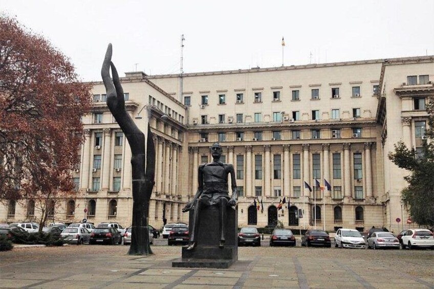 Une statue se trouve devant un grand bâtiment, orné de plusieurs drapeaux. On y voit également des voitures garées et un arbre aux feuilles d’automne. 3