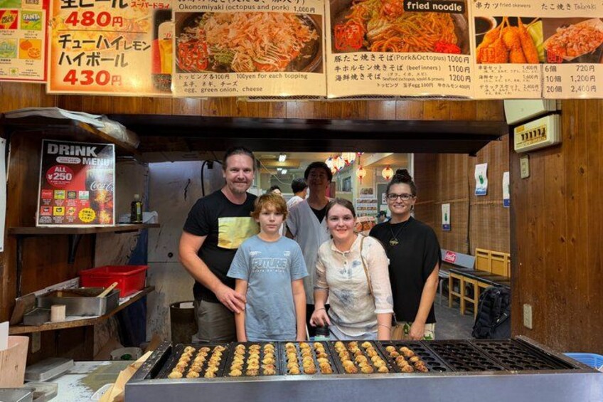 Una familia en un puesto de comida mostrando un menú con precios y platos de fideos fritos. 1