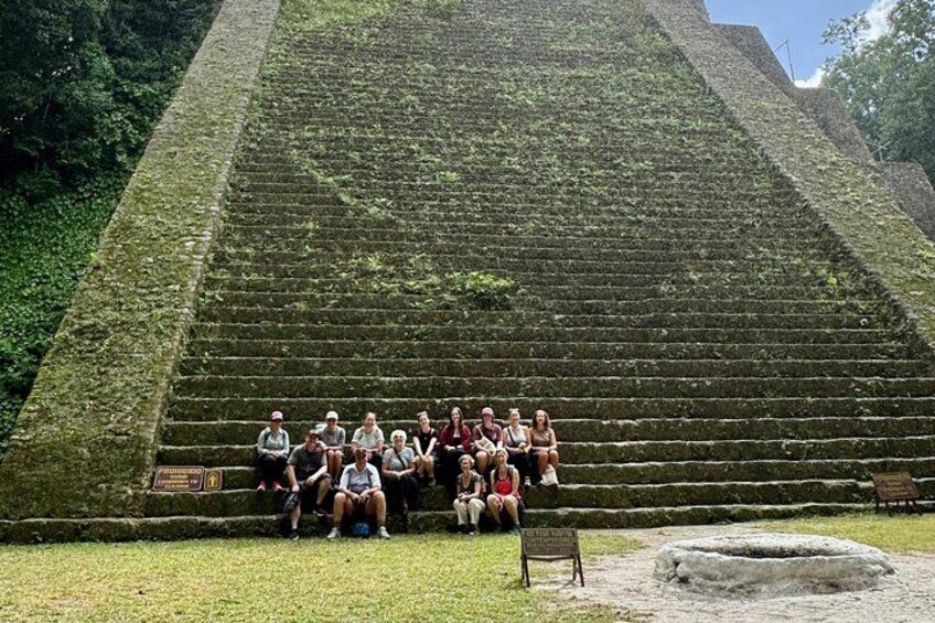 A group of people sitting on steps of a large pyramid-like structure surrounded by greenery. 4