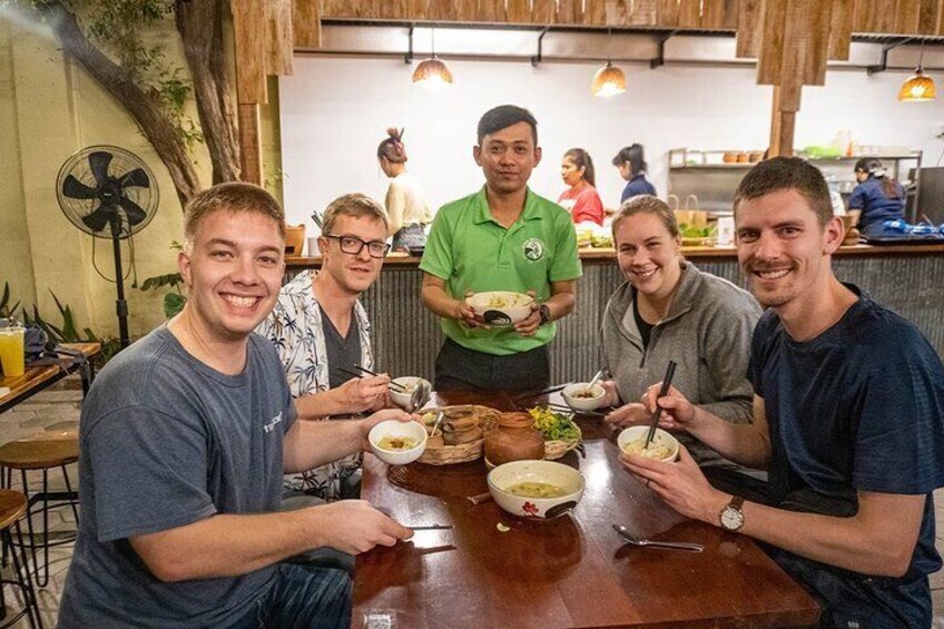 A group of people sitting at a wooden table in a restaurant, eating bowls of food. 1