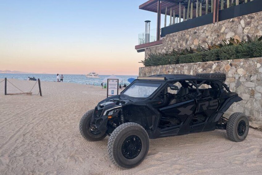 Un veicolo fuoristrada nero parcheggiato su una spiaggia sabbiosa, con un muro di pietra e un edificio sullo sfondo. 1