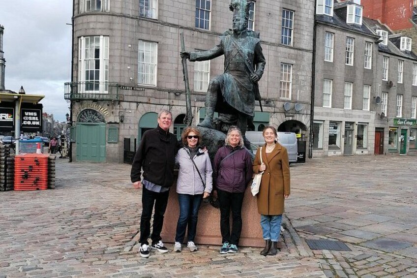 En grupp människor som står framför en staty på ett torg med kullerstenar, med byggnader i bakgrunden. 3