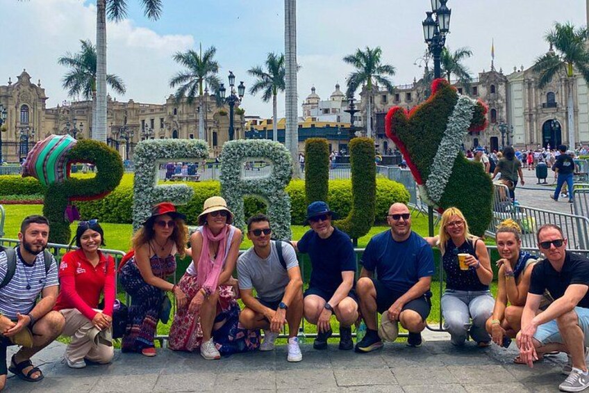 Een groep mensen poseert voor een park met grote letterbeelden en een historisch gebouw op de achtergrond. 1