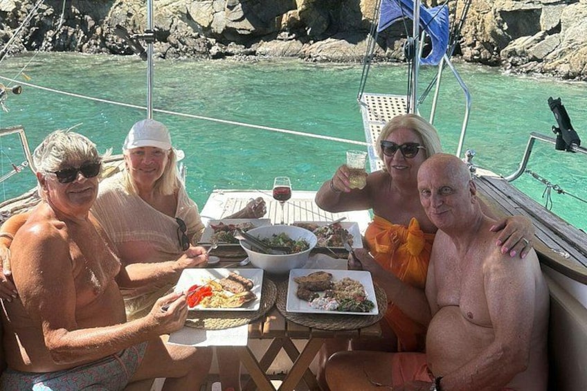 Un groupe de personnes est réuni autour d’un repas sur un bateau, avec une côte rocheuse en arrière-plan. 3