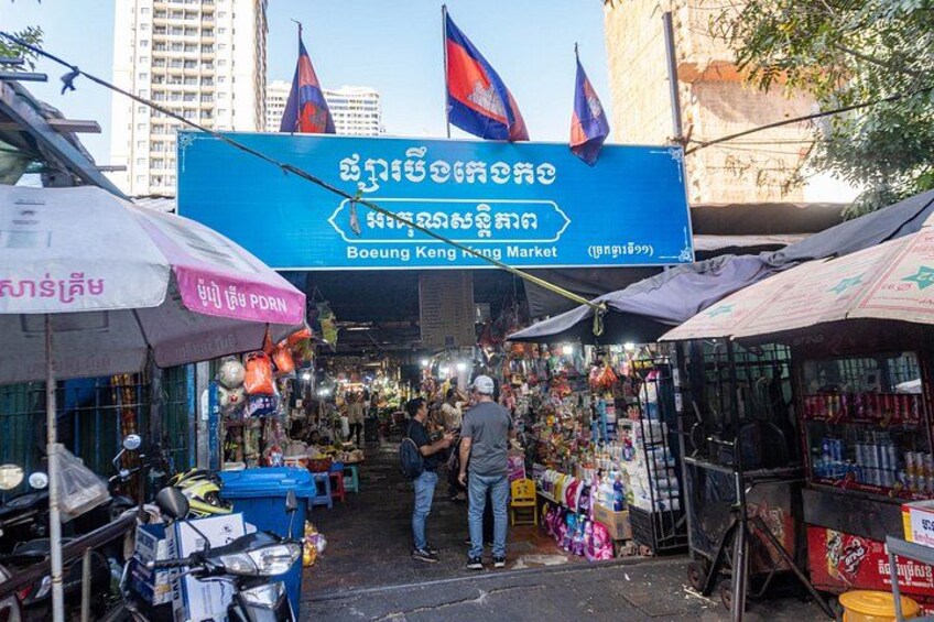 Un marché animé, reconnaissable à son grand panneau bleu, avec de nombreux étals et un homme debout au centre. 3
