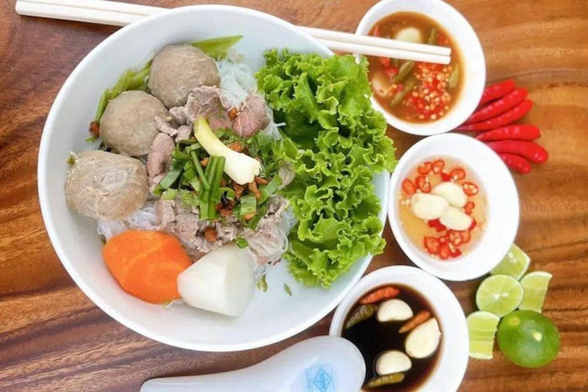 A bowl of Vietnamese noodle soup with meatballs, lettuce, and various condiments. 1