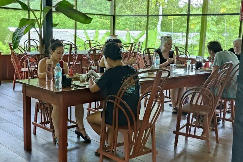 A group of people dining at a wooden table in a restaurant with large windows and plants. 1