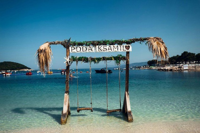 Een houten schommel met een bord waarop 'PODA KSAMIL' staat en een palmboom decoratie. 5