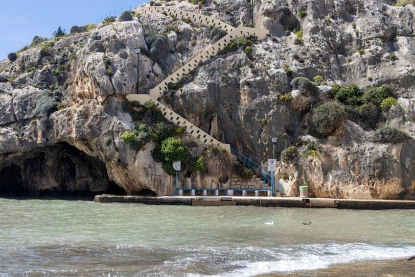 A rocky coastline with a staircase leading up to a cliff, a body of water, and a small structure with a sign. 1