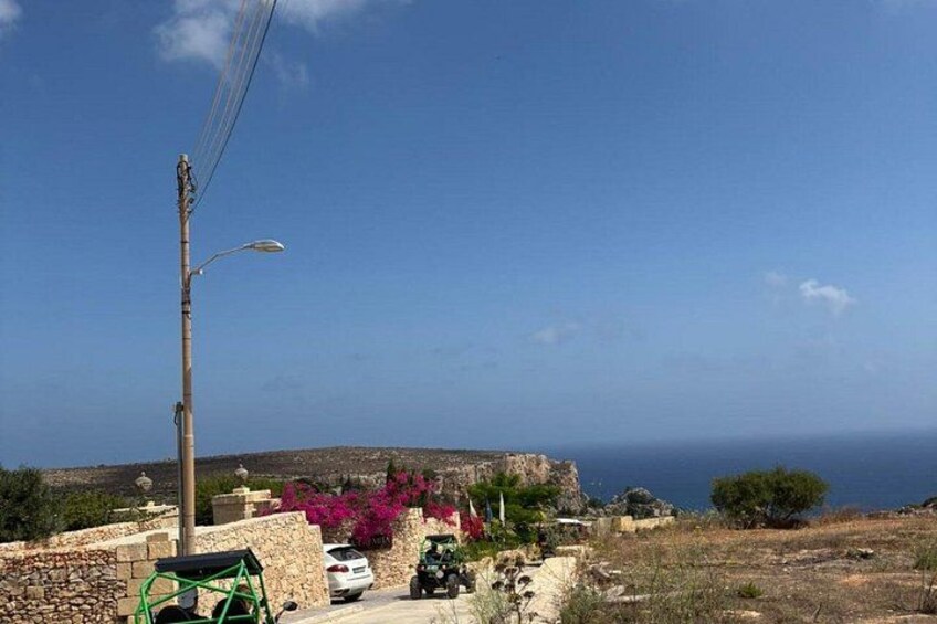 A coastal landscape with a stone wall, a street lamp, a parked car, and a row of pink bougainvillea flowers. 3