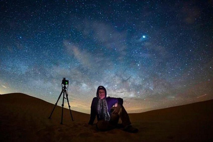 A person sitting on the ground under a starry night sky with a tripod-mounted camera. 2