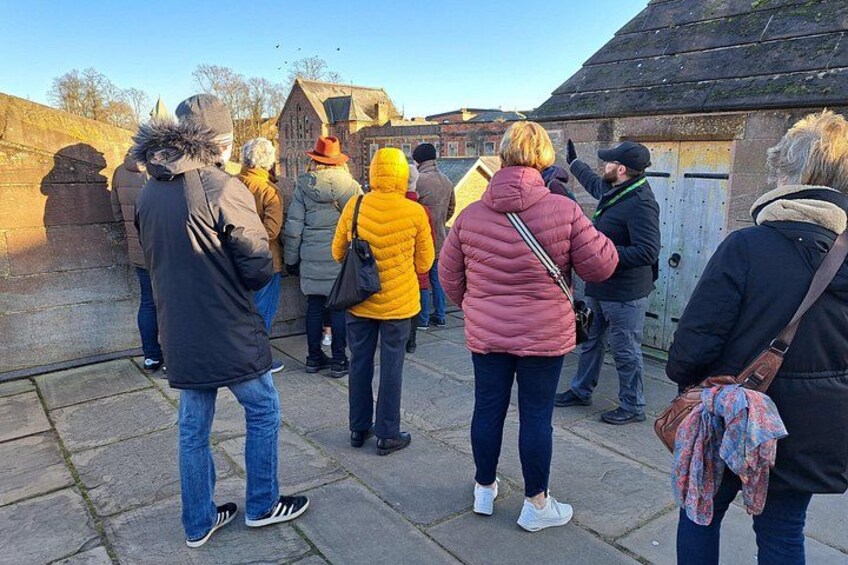 A group of people standing outdoors, some wearing winter jackets, with a stone wall and a building in the background. 1