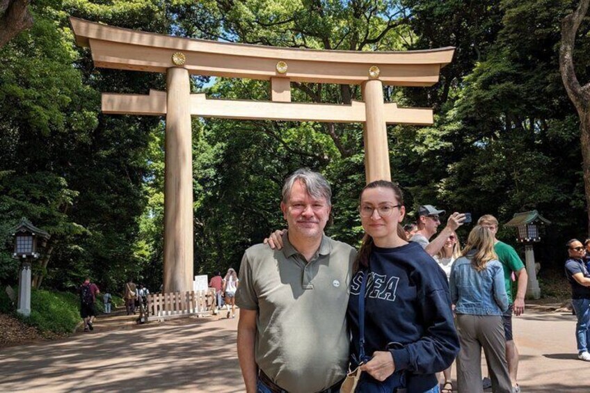 Two people standing in front of a large wooden torii gate in a park with trees and a pathway. 4