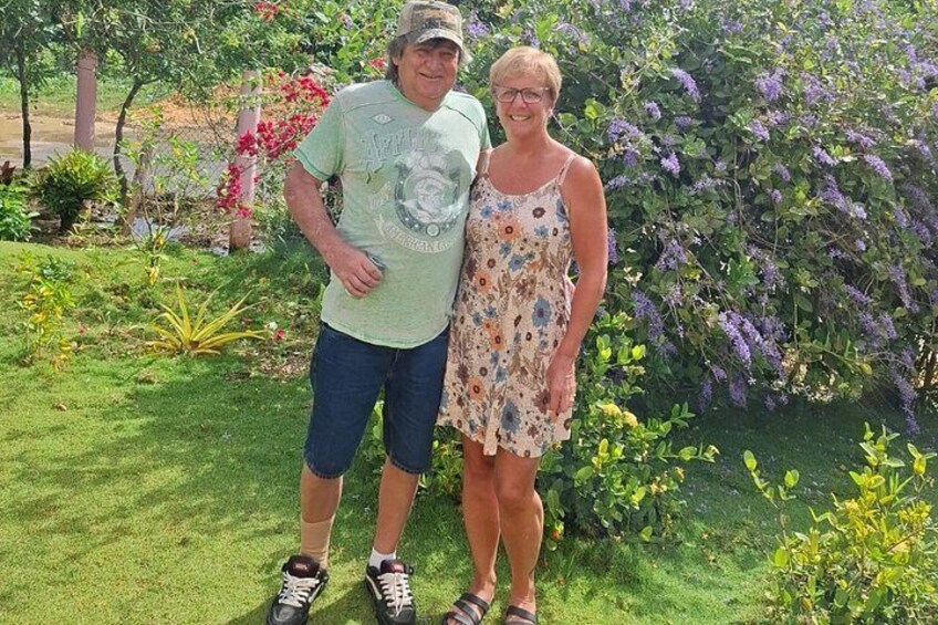 Two people standing in a garden with green grass, various plants, and a purple flowering bush. 5