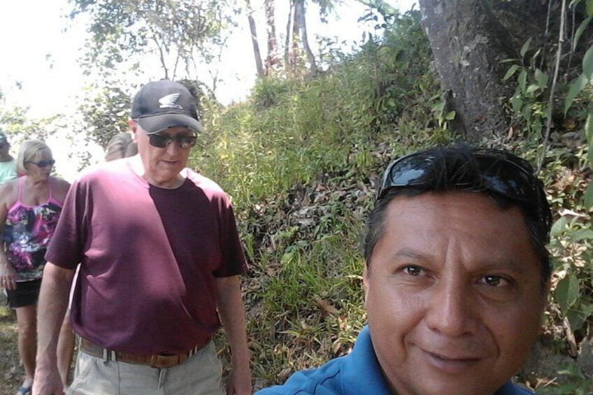 A person wearing a maroon shirt and a cap, standing outdoors with greenery and trees in the background. 3