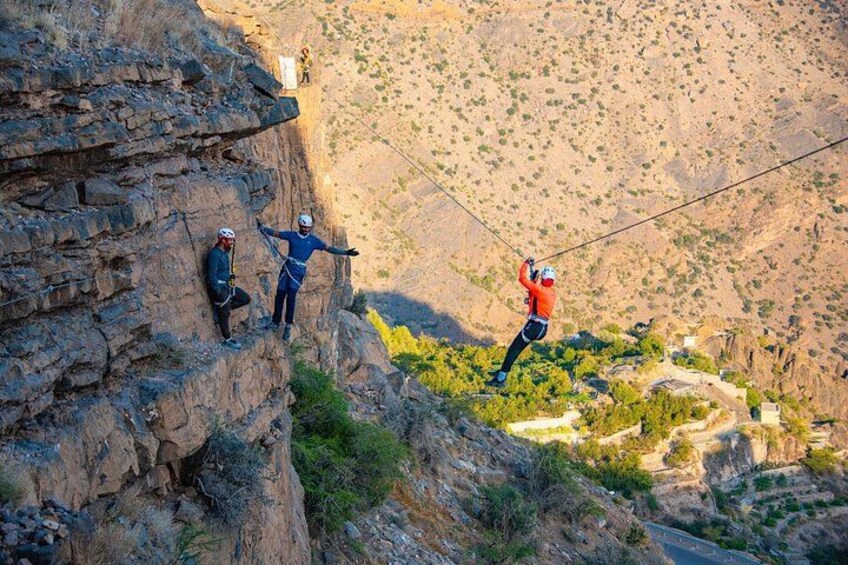 Three people on a mountain ridge, one standing on a cliff edge, another on a ledge, and the third on a rock face. 1