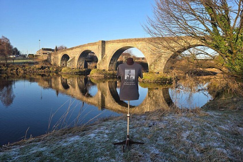 A stone bridge with three arches over a calm river, surrounded by grassy banks and bare trees. 3