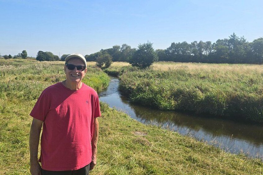 A man in a red shirt standing by a narrow water channel in a grassy field. 5