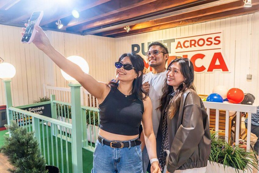 A person taking a selfie with a smartphone, a wooden wall with the text 'PUTT ACROSS AMERICA', and a green fence. 5