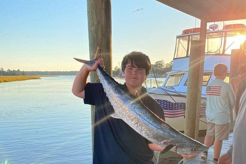 A person holding a large fish near a dock with a boat and American flag in the background. 3