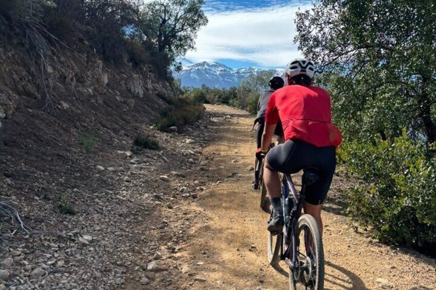 Two cyclists on a dirt path with rocky terrain and sparse vegetation, surrounded by hills and a clear sky. 2