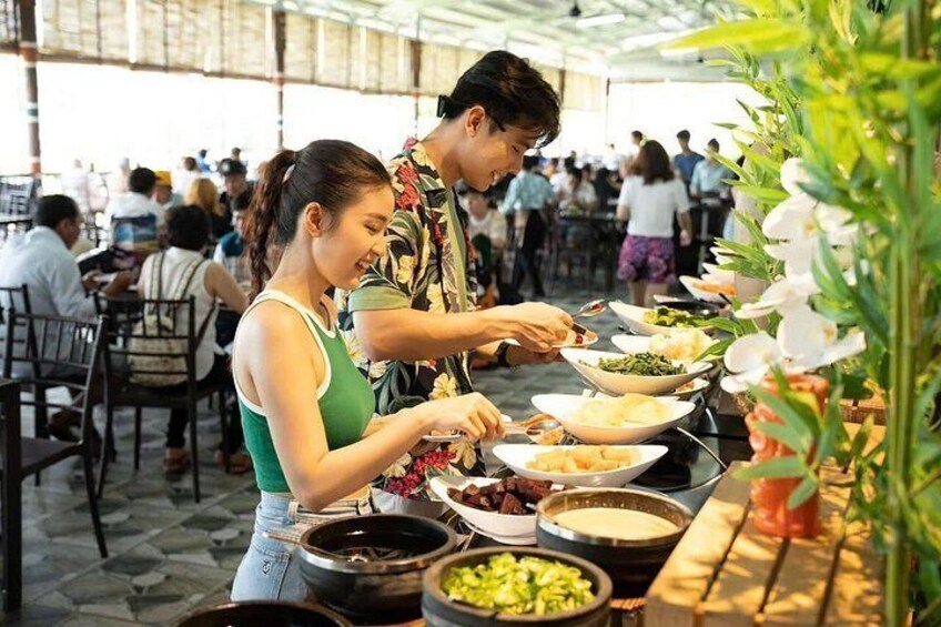 A group of people dining outdoors at a buffet with various dishes and a tropical plant in the foreground. 5