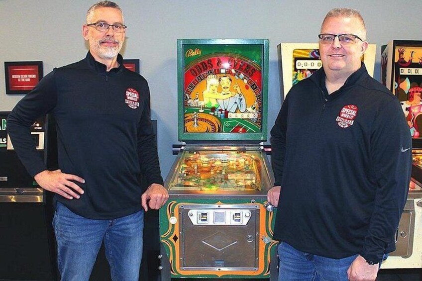 Dos personas de pie junto a una máquina recreativa de pinball antigua con una exhibición colorida y un compartimento para monedas. 5