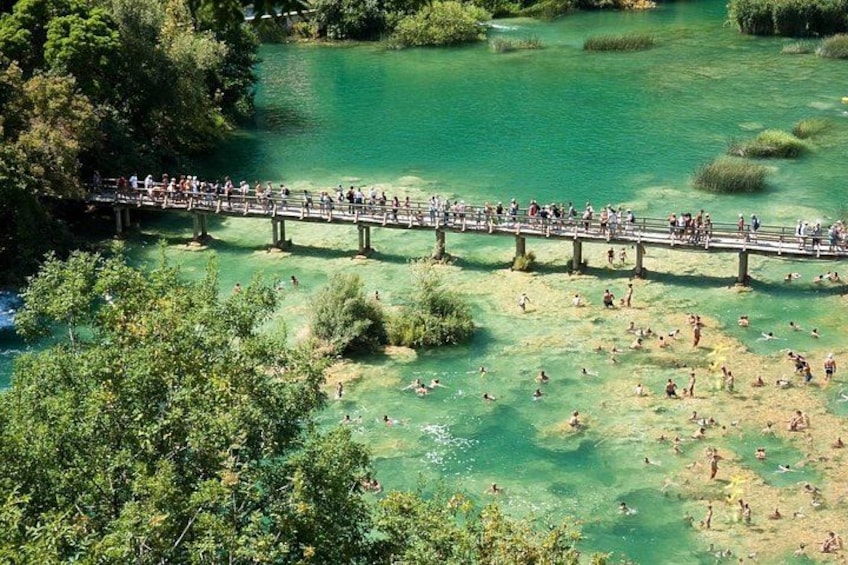 A bridge over a clear green lake with people swimming and walking. 2
