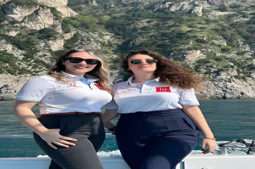 Dos personas con camisas blancas y distintivos rojos, de pie en un barco con una costa rocosa al fondo. 3