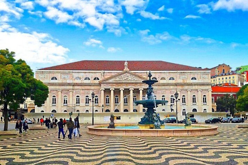 A public square with a fountain, a large building with classical architecture, and a patterned pavement. 2