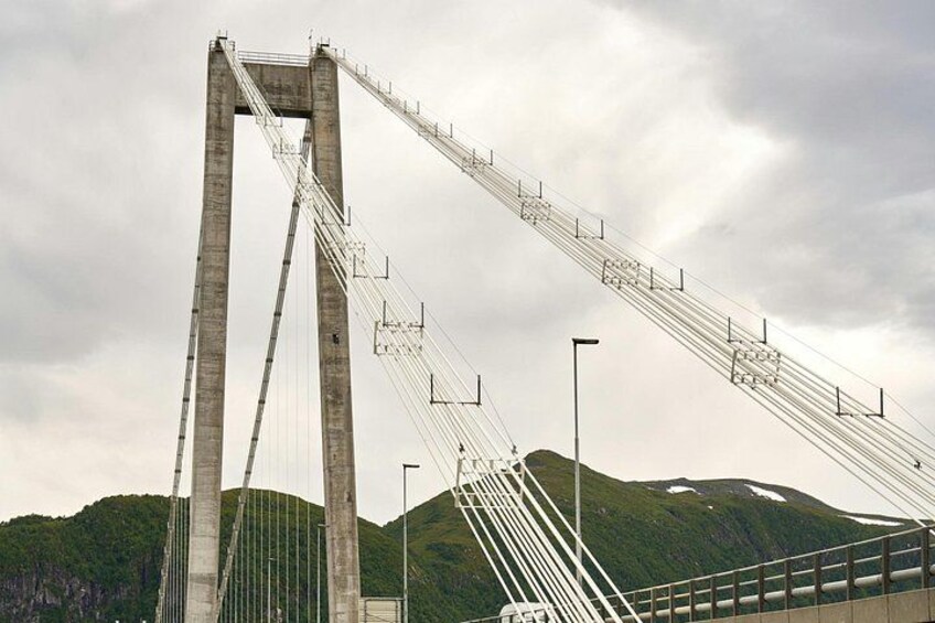 A large suspension bridge with tall towers and cables, set against a backdrop of green hills and a cloudy sky. 2