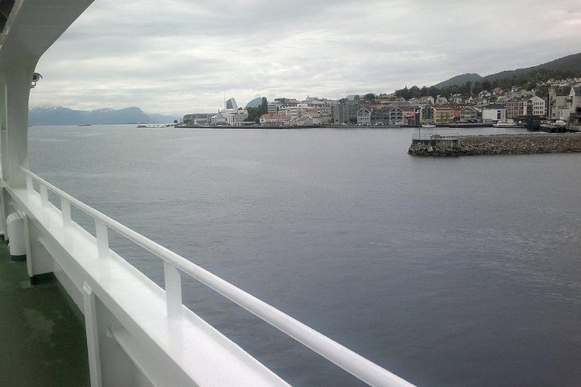 A calm body of water with a white railing, a distant shoreline with buildings, and mountains in the background. 5