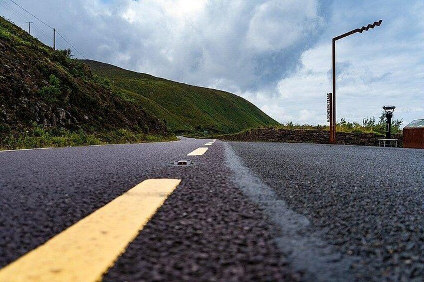 A road with yellow centerline markings, a guardrail on the left, and a hill in the background. 4