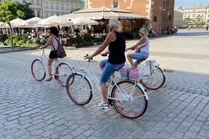 Three people riding bicycles in a public square with brick pavement and outdoor seating areas in the background. 2