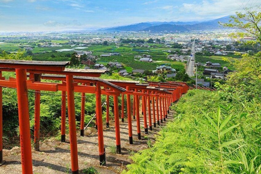 A row of red torii gates leading down a hill with a view of a town and mountains in the distance. 5