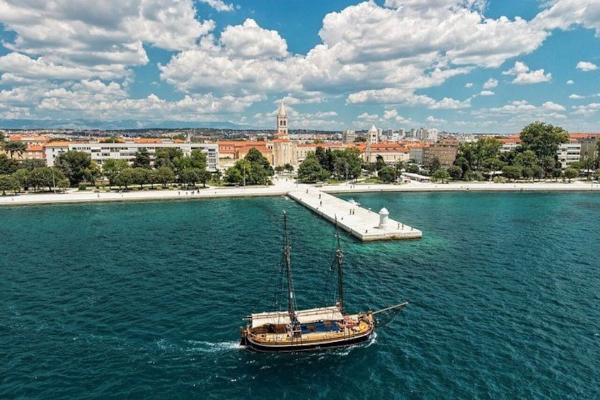 Een boot op een heldere blauwe zee, een steiger, en een stadsgezicht met een opvallende toren op de achtergrond. 2
