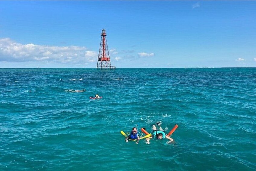 A lighthouse in the distance, people swimming and floating in the ocean, and a clear blue sky. 2