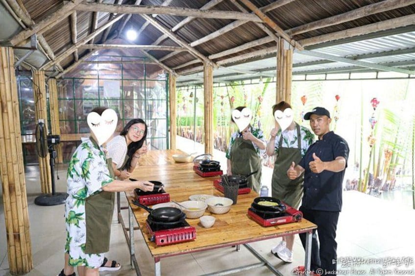 A group of people around a table with cooking equipment, in a covered outdoor area with bamboo walls. 5