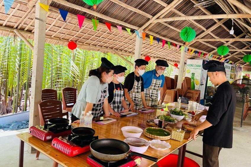 A group of chefs preparing food at an outdoor market with a thatched roof and palm trees in the background. 1