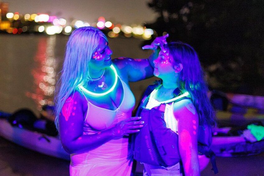 Deux personnes dont le corps est illuminé par des lumières vives, debout près d’une étendue d’eau, avec des bateaux en arrière-plan. 5