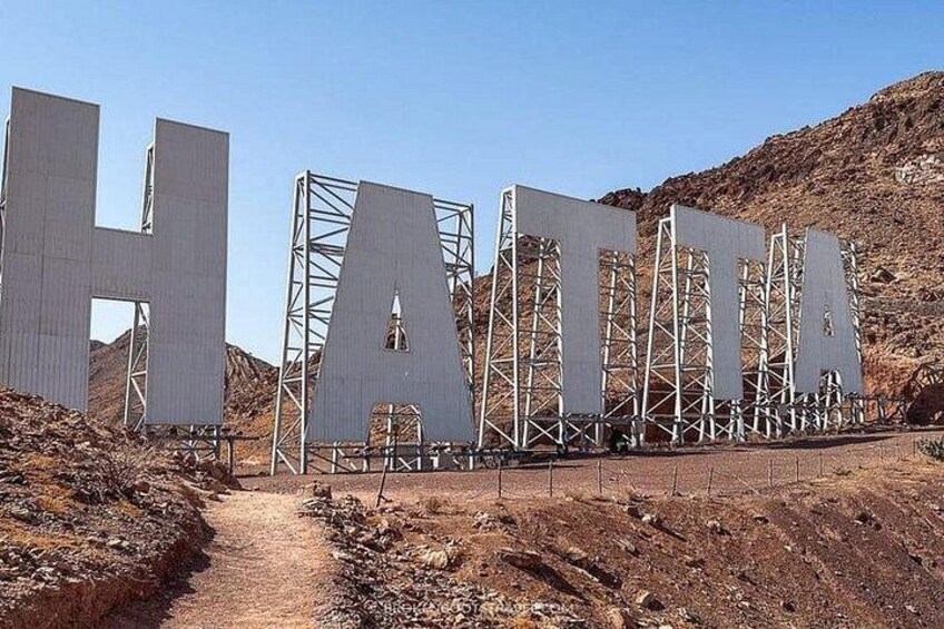 Large metal letters spelling 'HATTA' in a desert landscape with rocky terrain and hills in the background. 3
