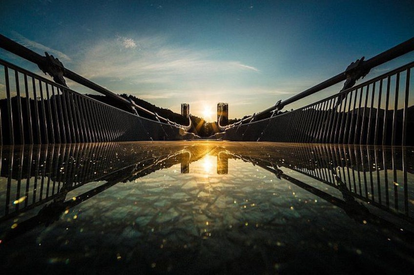 Un puente con piso de vidrio, barandas metálicas y una puesta de sol al fondo. 1