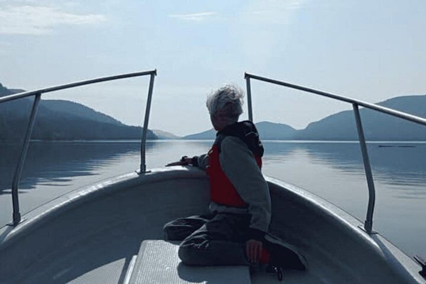 A person sitting on a boat with a metal railing, looking out over calm water with mountains in the distance. 2