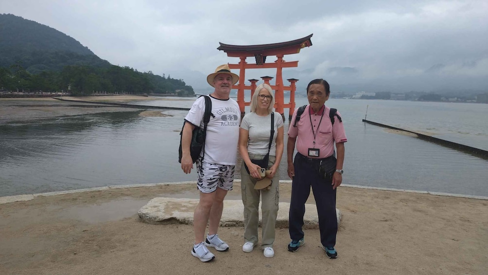 Three people standing in front of a red torii gate with a body of water and mountains in the background. 4