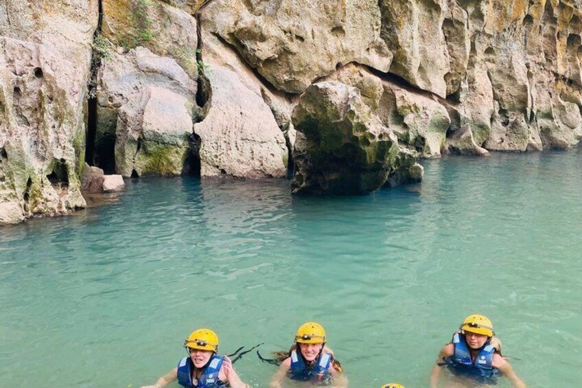 Three people in blue life vests and yellow helmets swimming in a turquoise body of water with rocky cliffs in the background. 4