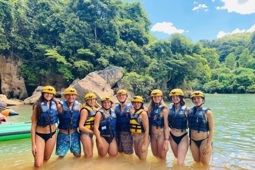 A group of people in water with helmets and life vests, surrounded by greenery and rocks. 2