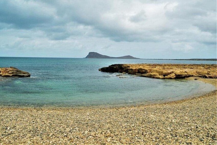 A pebble beach with calm water, distant rocky island, and cloudy sky. 4