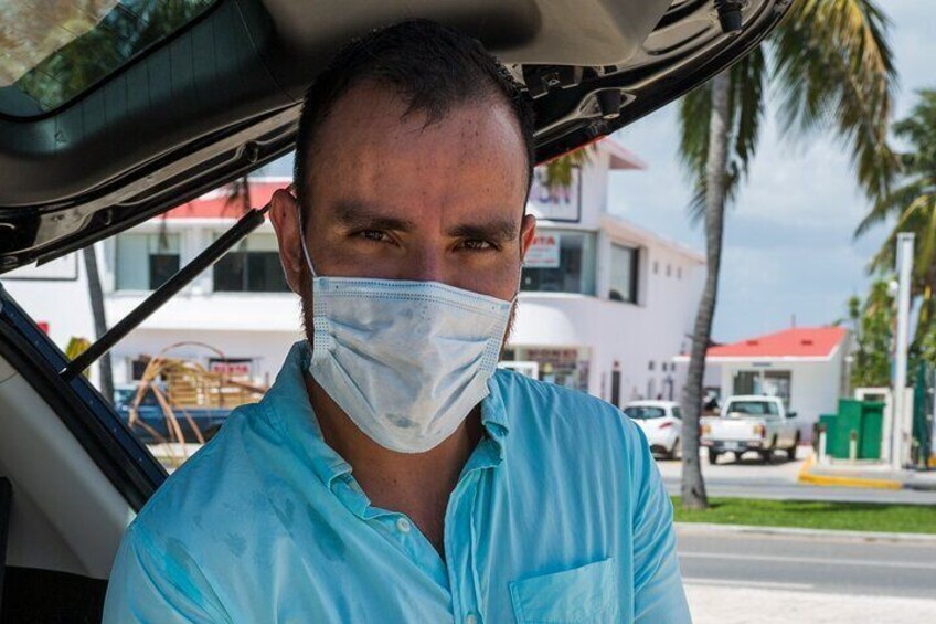 A person wearing a blue shirt sitting in a car with a building and palm trees in the background. 2
